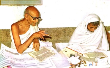 Jambuvijayji Maharaj Saheb in white robes is at a table, collaboratively analyzing papers.